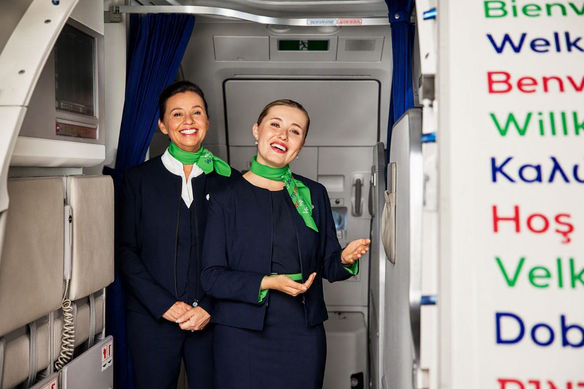Zwei lächelnde Flugbegleiterinnen in dunkelblauen Uniformen mit grünen Halstüchern stehen im Eingangsbereich eines Flugzeugs vor einer mehrsprachigen Tafel.
