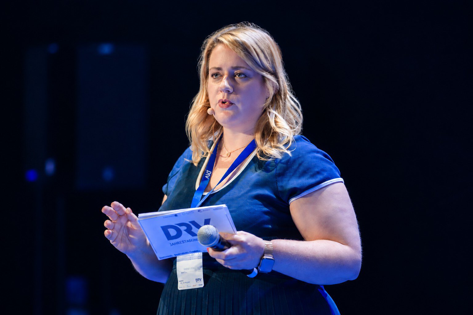 Eine blonde Frau mit blauem Kleid spricht vor einem dunklen Hintergrund in ein Mikrofon und hält eine Karte mit dem DRV-Logo in der Hand auf einer Bühne.