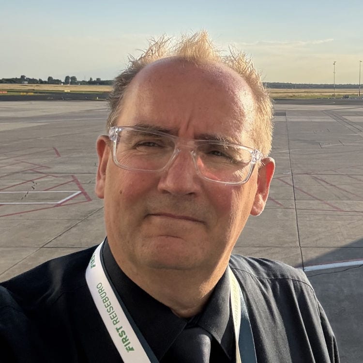 Ein Mann mit Brille und dunklem Hemd trägt ein Schlüsselband mit der Aufschrift FIRST REISEBÜRO vor einem weitläufigen, asphaltierten Flugfeld bei Tageslicht.