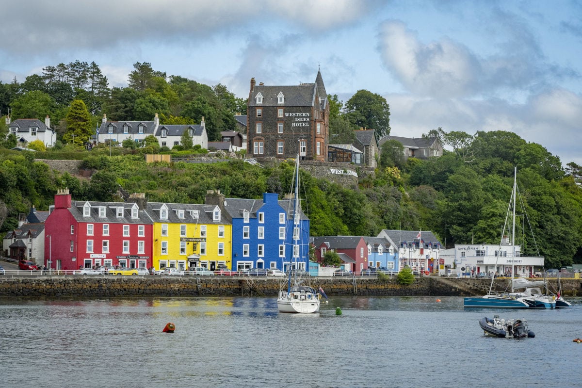 Blick auf die bunte Häuserzeile von Tobermory am Hafen mit dem Western Isles Hotel auf dem Hügel und verschiedenen Segelbooten im ruhigen Wasser davor.