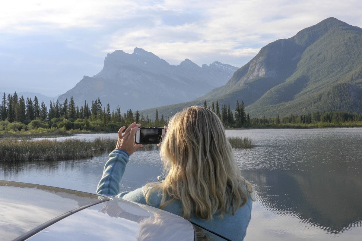 Eine blonde Frau in einem hellblauen Pullover fotografiert mit ihrem Smartphone eine Berglandschaft mit einem See und Nadelwald im Hintergrund.